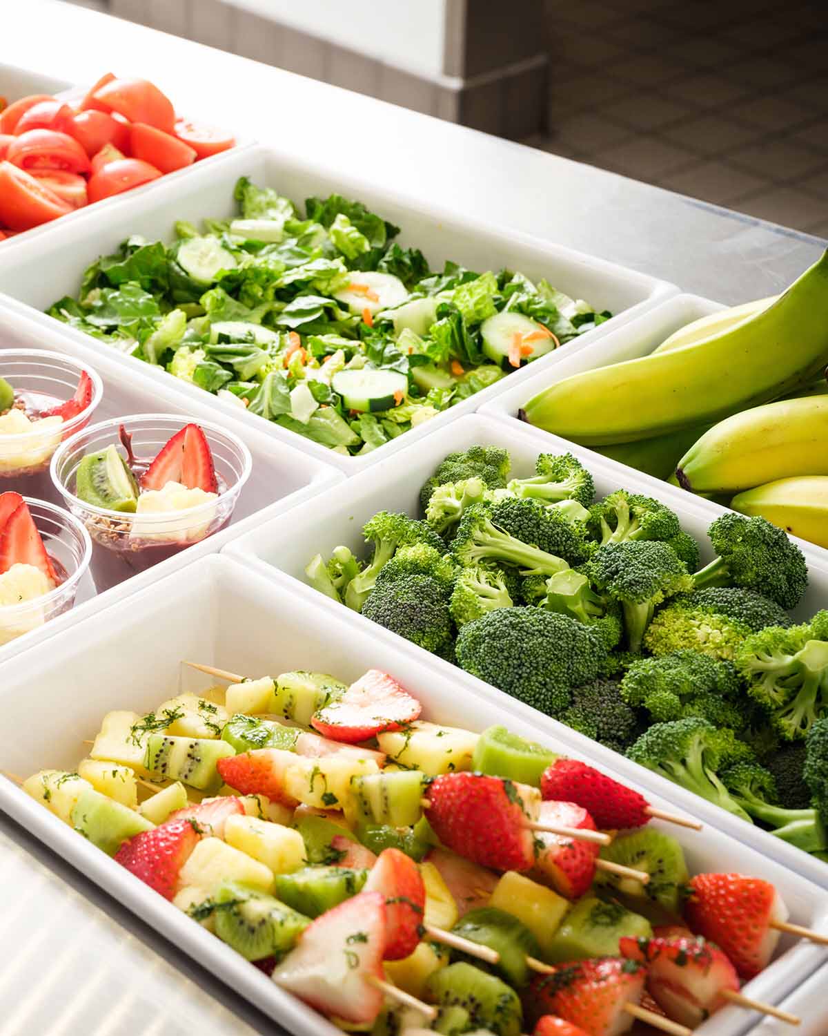 A variety of fresh vegetables, fruit cups, and bananas are neatly arranged on a cafeteria-style buffet.