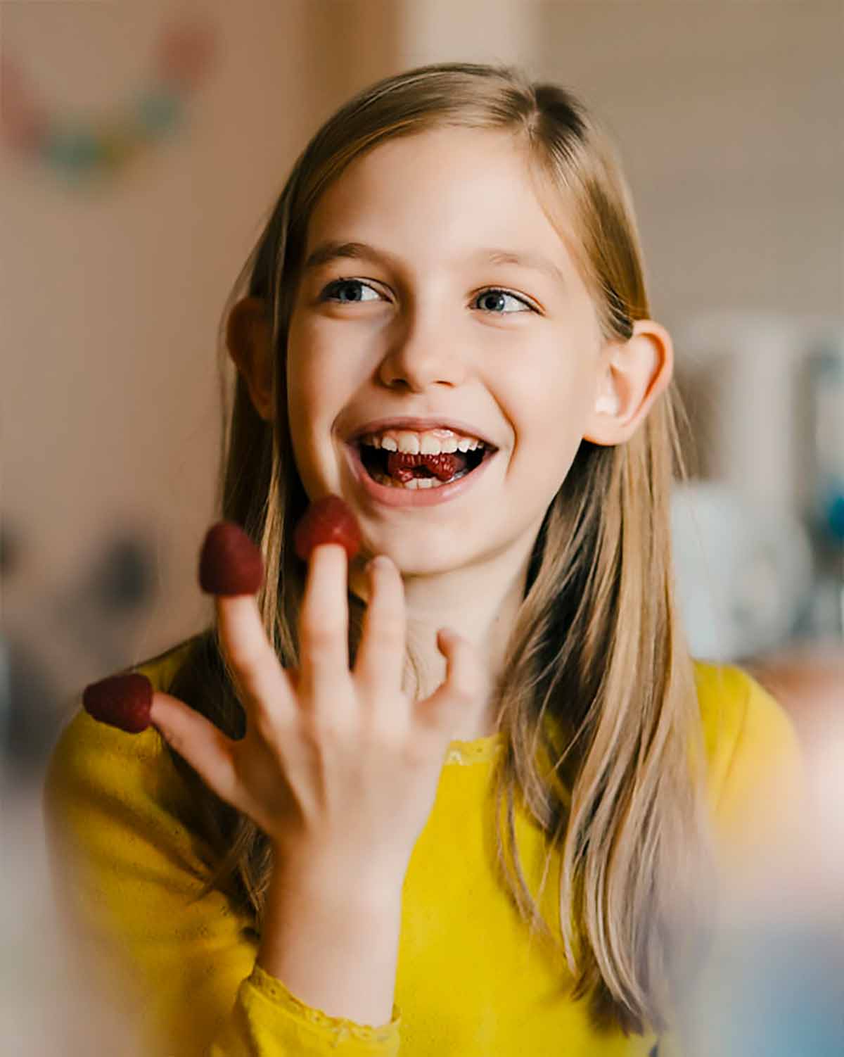 A cheerful young girl with long blonde hair wearing a yellow shirt, smiling with her fingers covered in chocolate in a cozy indoor setting.
