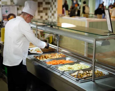 A chef dressed in white uniform and hat is serving food from a buffet-style hot food counter in a cafeteria setting.