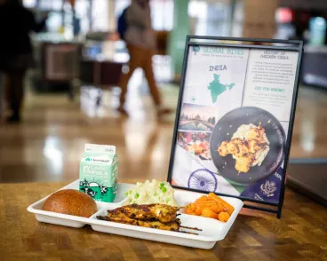A cafeteria-style tray with a meal of chicken tikka, steamed carrots, mashed potatoes, a bread roll, and a carton of milk, placed next to an informational sign about India and chicken tikka in a dining area.
