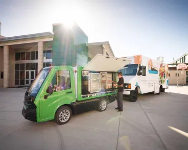 A worker serves from a compact green tuktuk food truck parked outside a high school by another food truck.