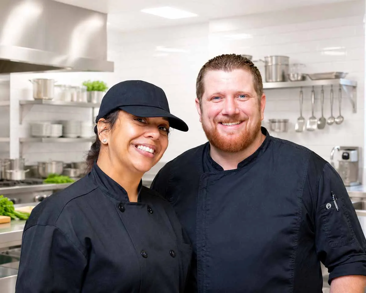 Two smiling chefs in black uniforms standing together in a kitchen or bakery setting.