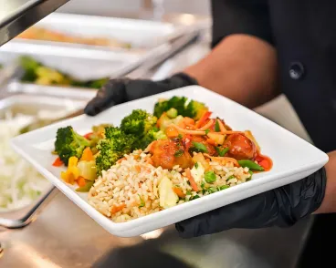 A person wearing black gloves holding a plate filled with fried rice, mixed vegetables, and broccoli in a buffet or cafeteria setting.