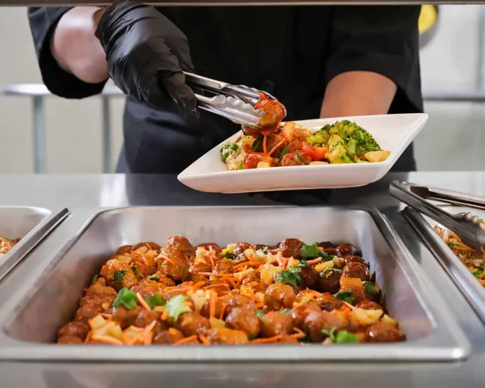 A person wearing black gloves is serving food with tongs onto a white plate from a buffet tray filled with meatballs and vegetables in a cafeteria or food service setting.