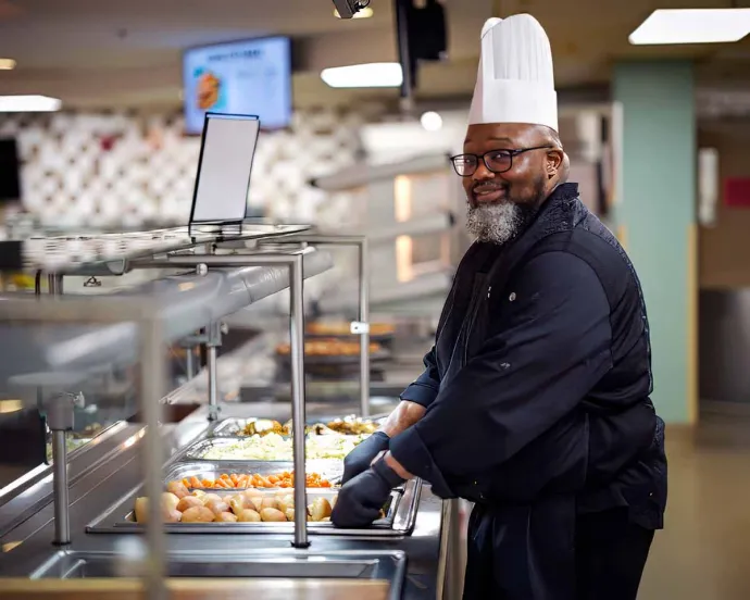 A chef wearing a white hat and black uniform is smiling while serving food in a cafeteria setting.