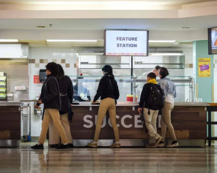 A group of students in standing in line at a cafeteria counter labeled "Feature Station" to get food.