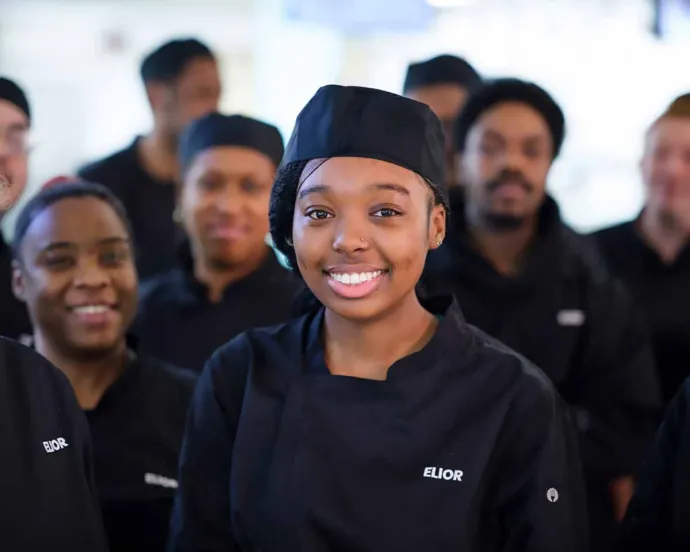 A group of smiling chefs, with a young woman in the center wearing a black chef uniform and hat, standing in a professional kitchen.