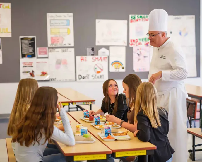 A chef in a white uniform talks to four girls sitting at a cafeteria table eating lunch, with posters on the wall behind.