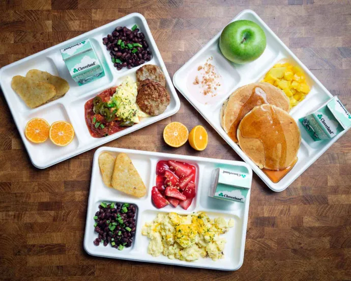 Three cafeteria trays on a wooden surface, each filled with different breakfast foods including pancakes, scrambled eggs, fruit, and milk cartons.