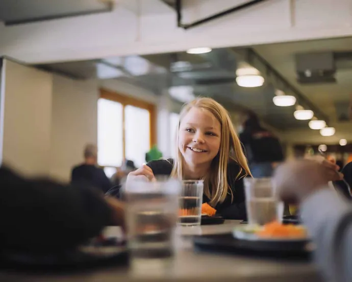 A young girl is smiling and eating a meal with others at a brightly lit indoor cafeteria.