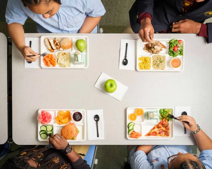 Four students are sitting around a cafeteria table, eating different meals from lunch trays in a school setting.