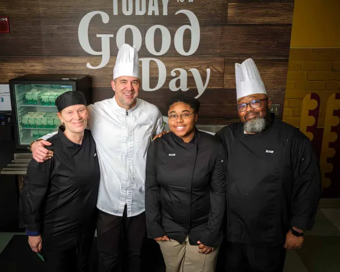Four chefs, two wearing white chef hats and two in black uniforms, standing closely together and smiling in a kitchen setting with a wooden backdrop that reads "Today is a Good Day."